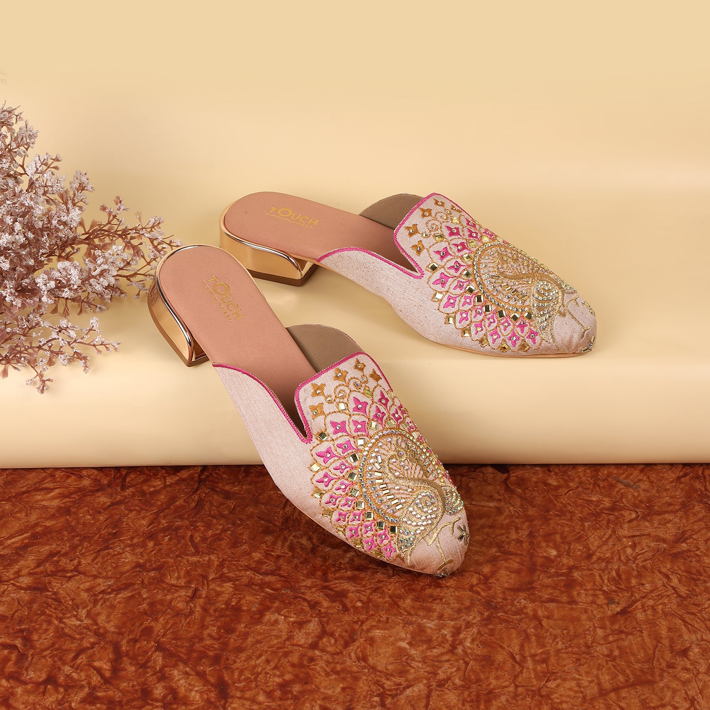 Elegant embroidered mule slippers with intricate peacock motif and sparkling rhinestones, showcased on a warm brown surface alongside a vibrant floral arrangement.